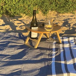 A wooden picnic table in natural tones on sand, holding a wine bottle and glass, with a checkered and striped blanket.