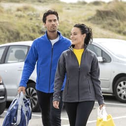 A man in a blue fleece jacket and a woman in a grey fleece jacket walking outdoors, both carrying bags.