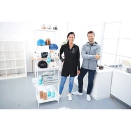 A man in a grey knit zip hoody and a woman in a black shirt stand by a display of products, including caps and drink bottles.