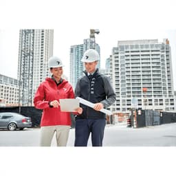 A woman in a red rain jacket and a man in a black jacket stand outside at a construction site, both wearing hard hats.