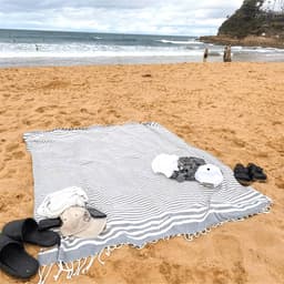 A striped throw blanket in grey and white lies on sandy beach, with a hat and sandals nearby.