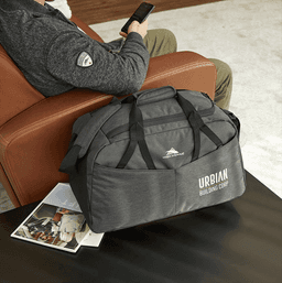 A grey duffel bag with multiple pockets and a logo, resting on a table next to a person sitting on a couch.