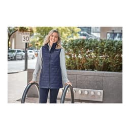 A woman wearing a navy insulated puffer vest over a grey top, standing outdoors with greenery in the background.