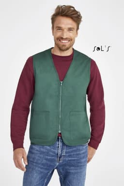 A green unisex zippered vest with two pockets, worn over a maroon long-sleeve shirt and denim jeans. It has a logo.