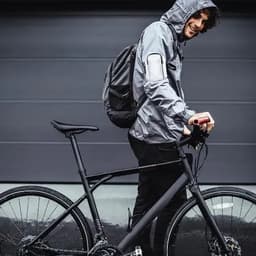 A man in a grey hooded jacket stands next to a black bicycle, holding the handlebars while smiling.