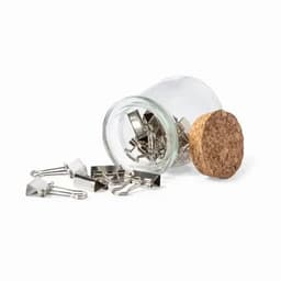 A glass jar with a cork lid filled with silver binder clips, some spilled onto a white surface.