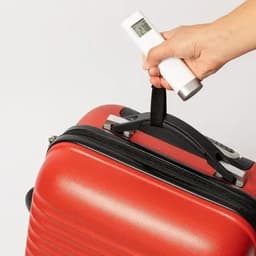 A hand holds a white luggage scale attached to a red suitcase with a black handle.
