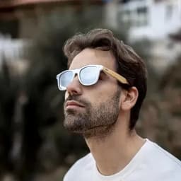 A man wearing white sunglasses with wooden arms, looking upwards against a blurred background.