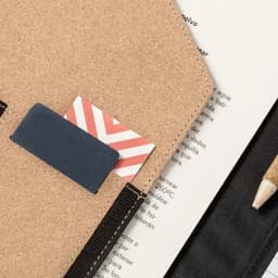 A cork notebook cover with two cards peeking out, alongside a pencil and printed pages.