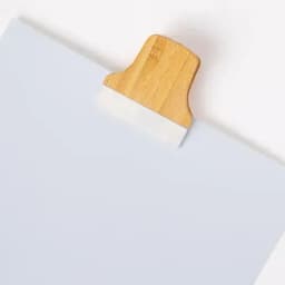 A clipboard with a wooden clip holds a light grey sheet of paper, featuring a simple and clean design.