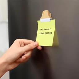 A hand holding a bright yellow sticky note attached to a clip on a metallic surface. The note contains handwritten reminders.