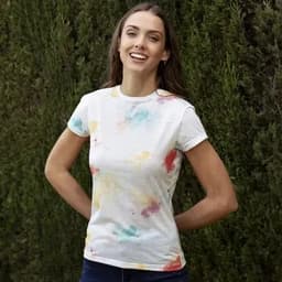 A woman in a white t-shirt featuring a vibrant, multi-coloured splatter design, standing outdoors against greenery.