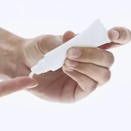 A hand holding a small white tube, dispensing its contents onto a fingertip. The tube has a plain design.