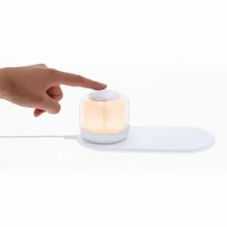 A light diffuser with a transparent dome and white base, emitting a warm glow, and being touched by a hand.