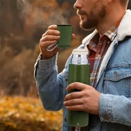 A drink bottle and a small green cup held by a man in a denim jacket, surrounded by autumn foliage.