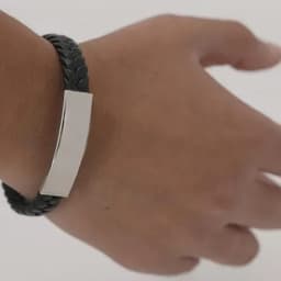 A black braided bracelet with a silver rectangular charm, worn on a wrist.