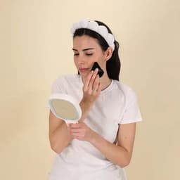 A woman in a white t-shirt holds a hand mirror and applies makeup with a black sponge, wearing a white headband.