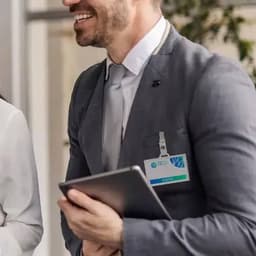 A man in a grey suit holds a tablet, smiling, with a name badge attached to his lapel.