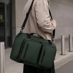 A green backpack with multiple zippered pockets, carried by a person wearing a beige jacket.