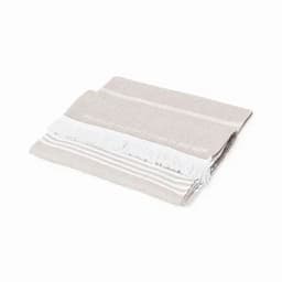 A folded towel in light beige and grey, featuring white fringes and decorative stripes. It has a subtle branding logo.