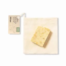 A solid soap bar in a natural cream colour, packaged in a small fabric bag. The soap includes visible exfoliating particles.