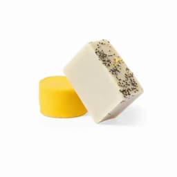 Two soap bars are displayed: one is yellow and round, and the other is rectangular with a beige surface sprinkled with black seeds.