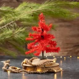 A small red Christmas tree made of synthetic bristles, placed on a burlap-covered wooden base, surrounded by greenery.