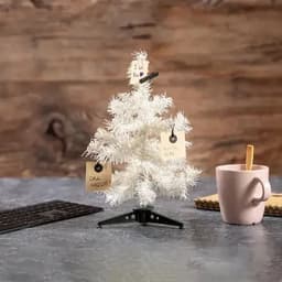 A small white artificial Christmas tree with tags and a pink mug beside it on a wooden table.