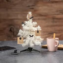 A decorative white Christmas tree with brown tags and clips, beside a pink mug on a wooden table.