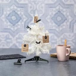 A small white artificial Christmas tree with tags attached, placed beside a pink mug on a grey surface.