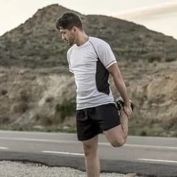 A man in a grey and black athletic outfit stretches his leg beside a road, with a rocky hillside in the background.