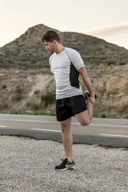 A man stretches while wearing black sports shorts and a grey athletic top, set against a rugged landscape.