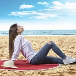 A woman relaxing on a beach towel, wearing a white top and jeans, with a straw hat beside her on a sandy beach.