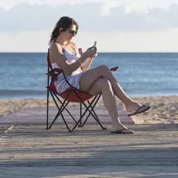 A woman sitting on a beach, wearing sunglasses and a striped outfit, using a phone while seated in a red beach chair.
