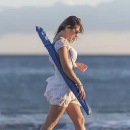 A woman in a white dress walks along the beach, holding a blue umbrella. The ocean waves are in the background.