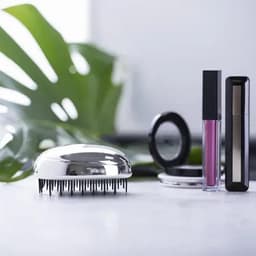 A shiny hairbrush with long bristles next to a compact mirror and lip products against a green foliage backdrop.