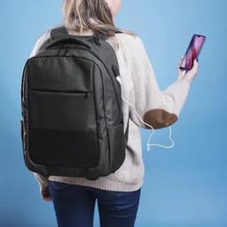 A backpack in charcoal grey with a USB charging port, worn by a person holding a smartphone on a blue background.