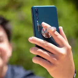 A smartphone with a blue back, featuring a ring grip on the rear, held by a person against a green background.