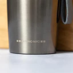 A drink bottle in stainless steel with a smooth finish and a grey strap, featuring a logo at the base.