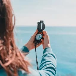 A compass with a black casing is held by a person against a backdrop of water and a clear sky.