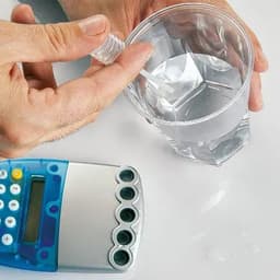 A person’s hand holds a small bottle above a clear cup of water, with a calculator placed nearby.