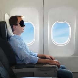 A person wearing a sleep mask sits by an airplane window, gazing peacefully outside. The interior features grey seating.