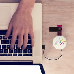 A hand typing on a laptop beside a pink and white promotional product with a logo. A cord connects it to the laptop.