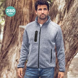 A man wearing a grey jacket with a zip and side pockets stands outdoors near trees. The jacket has a logo.
