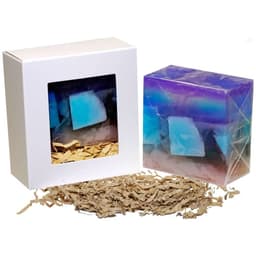 A soap bar in a box featuring blue and purple hues, with shredded paper filling the bottom. The box has a window.