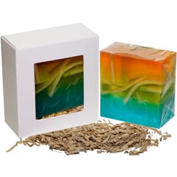 A colourful soap cube in orange and turquoise, presented in a white box with a clear window and shredded paper beneath.