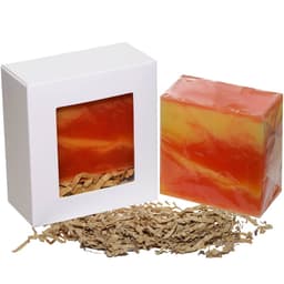 A soap product in an orange and yellow swirl, packaged in a white box with window, alongside brown shredded paper.