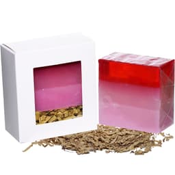 A pink and red layered soap bar beside a white box with a window, placed on decorative shredded paper.