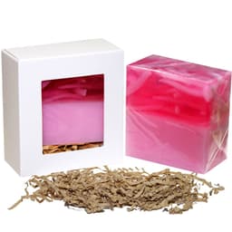 A pink and red soap bar in a clear box, with shredded brown paper underneath for packaging. The box has a logo.