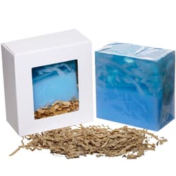A blue soap bar packaged in a white box with a clear window, accompanied by shredded paper for cushioning.